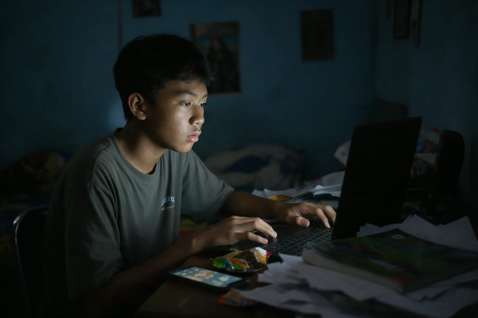 Seorang remaja laki-laki sendirian di meja kamar pada dini hari, wajahnya hanya diterangi cahaya layar laptop.