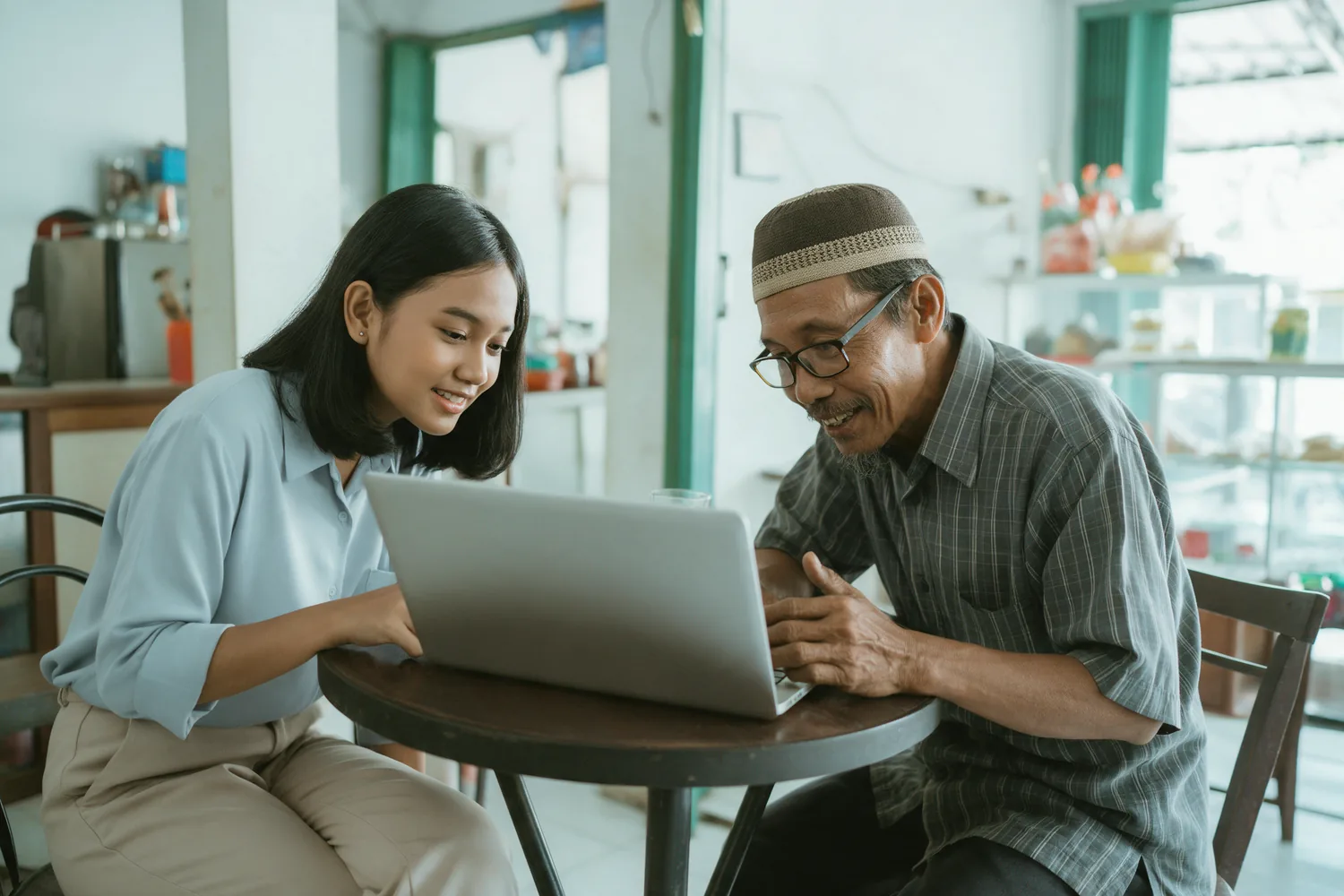 Seorang perempuan muda menunjukkan laptop kepada seorang pemilik warung yang lebih tua di meja kafe.
