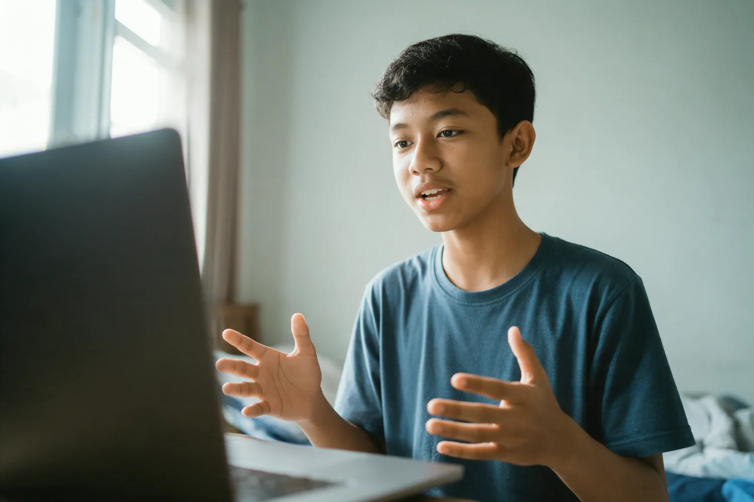 Seorang remaja laki-laki berbicara ke arah webcam laptopnya di kamar.