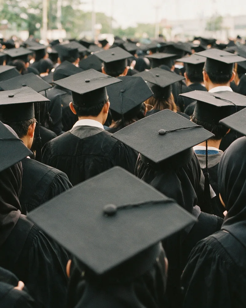 Deretan rapat wisudawan bertoga difoto dari belakang, topi demi topi memenuhi bingkai sampai jauh ke belakang.