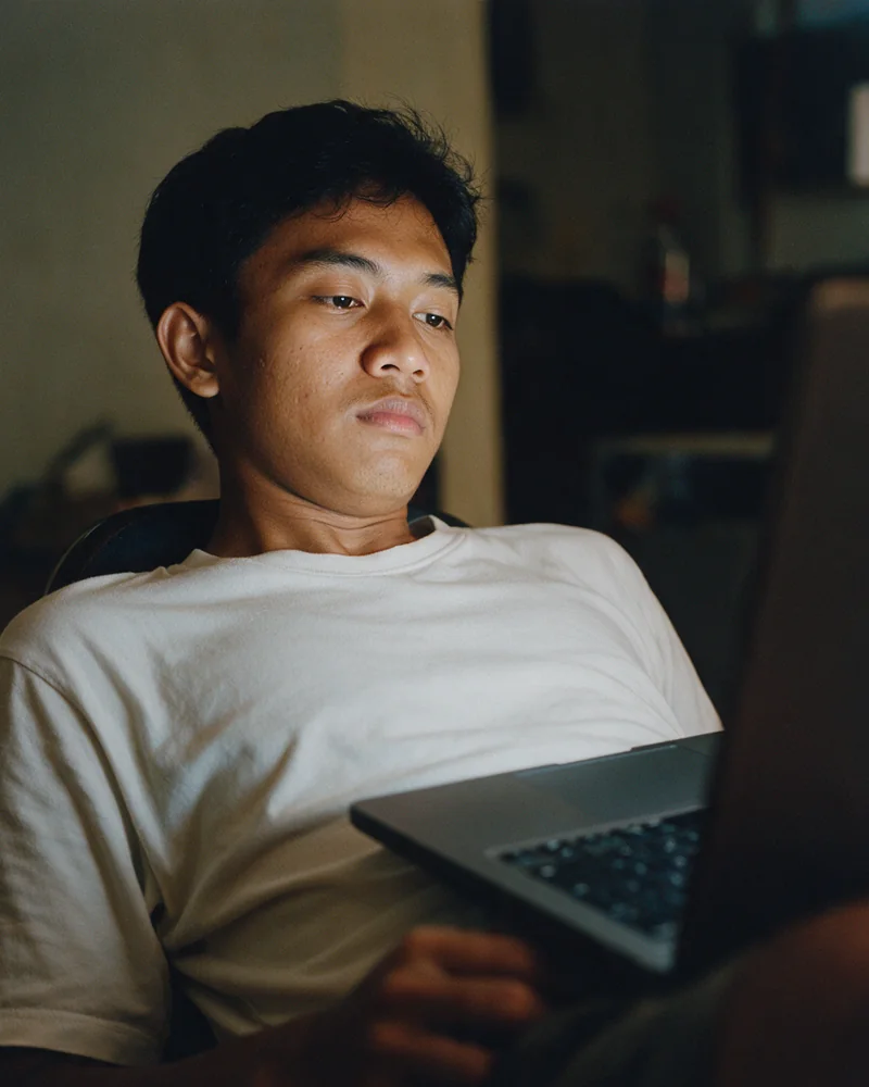 Seorang pemuda bersandar menjauh dari laptop yang terbuka, kedua tangannya lepas dari papan ketik, wajahnya diterangi cahaya layar.